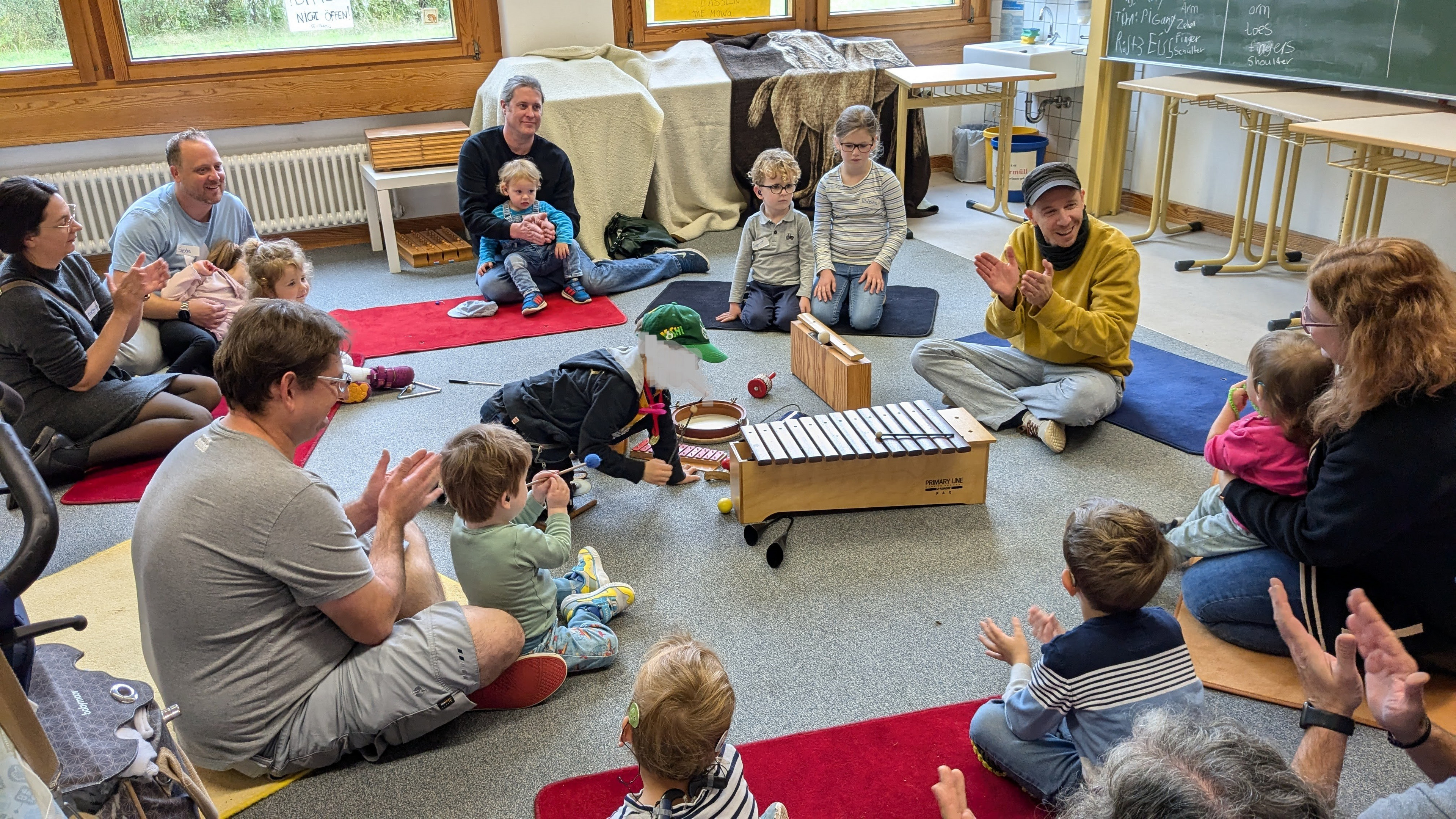 Eltern und Kleinkinder sitzen im Kreis auf dem Boden und musizieren mit Orff-Instrumenten.