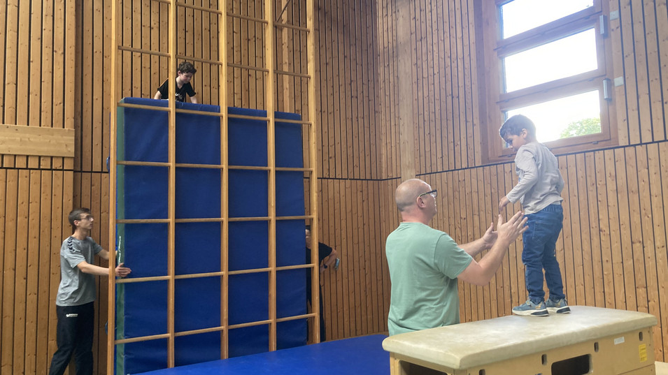 In der Turnhalle klettern Kinder auf einem Kasten und einem Klettergerät.