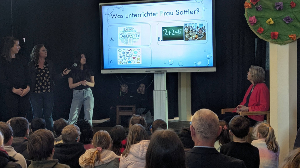 Schüler machen ein Rate-Quiz mit der Schulgemeinschaft und zeigen dazu die Fragen an einem Smartboard.