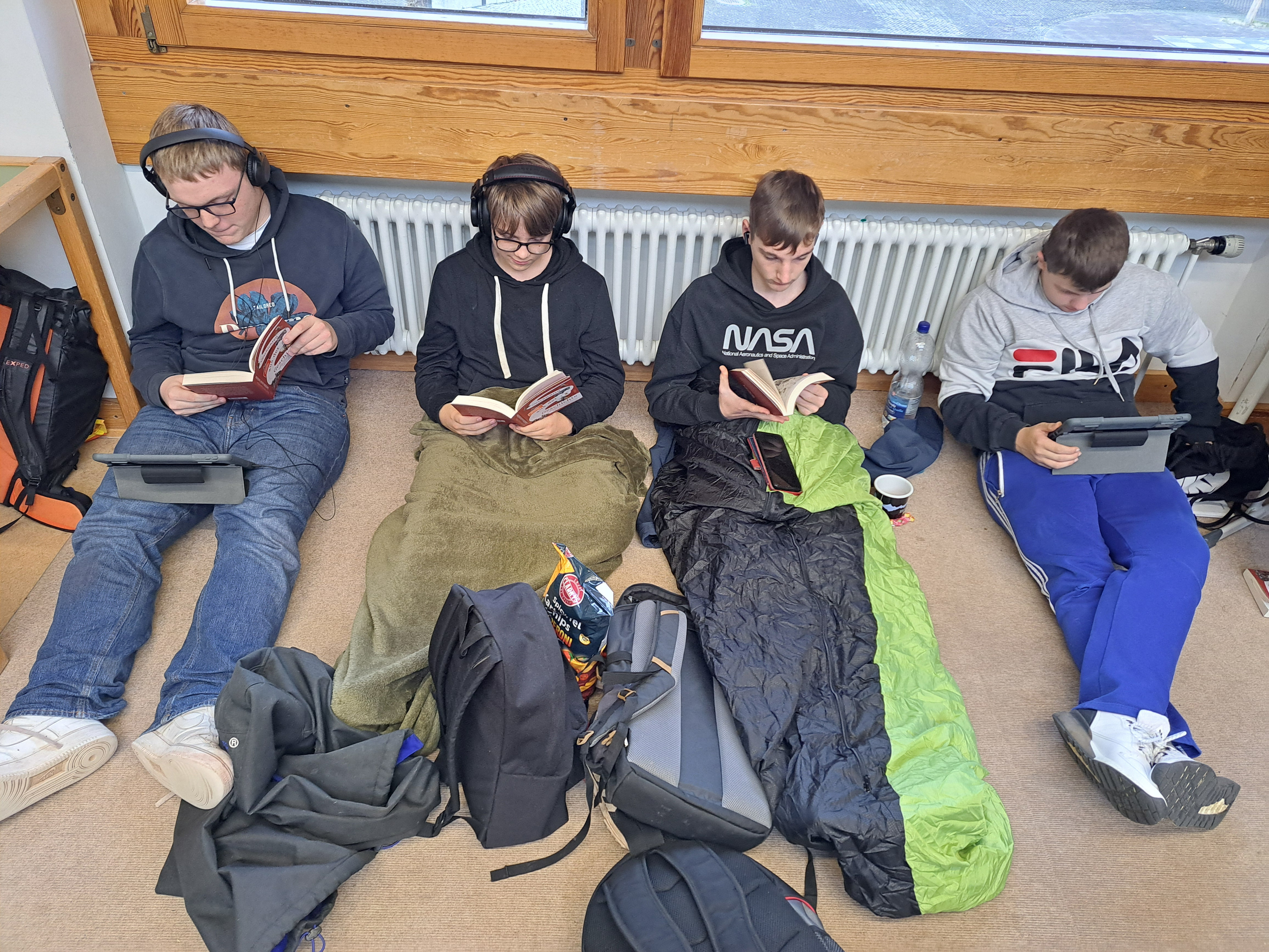 4 Schüler sitzend auf dem Klassenboden lesen in Bücher bzw. mit I-Pads. 
