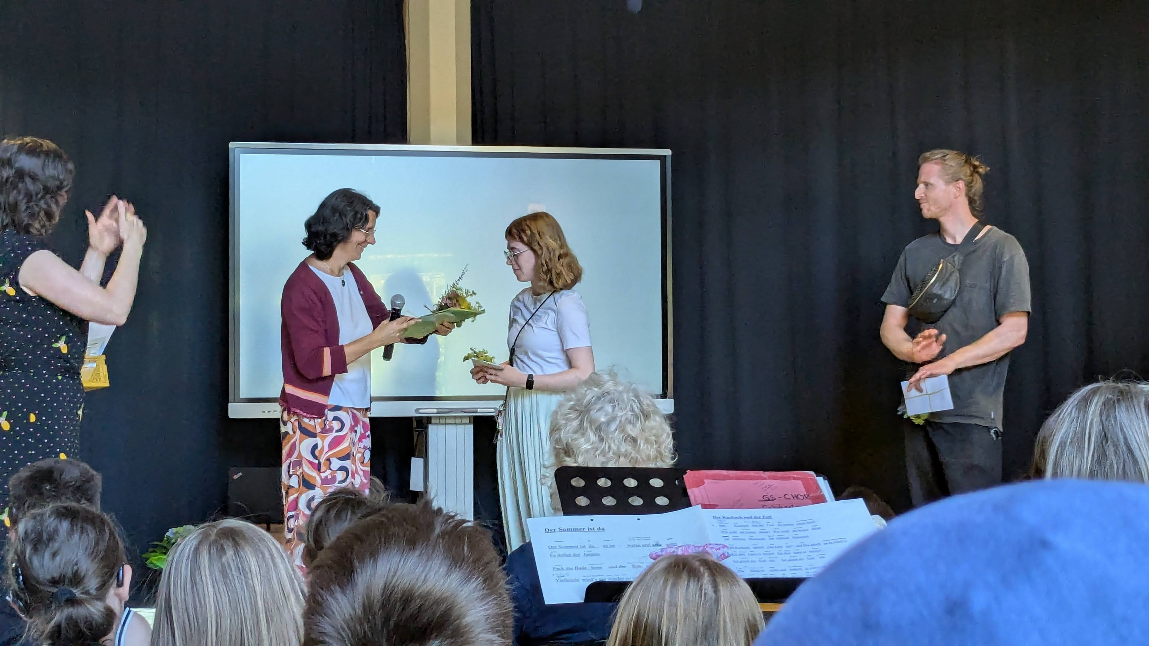 Eine Lehrkaft überreicht ein Geschenk an die Praktikantin und an den Lehramtsanwärter zum Abschied auf der Bühne