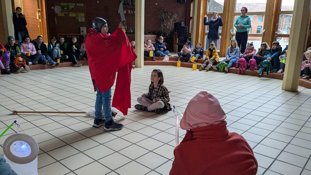 Schüler als St. Martin verkleidet mit Steckenpferd steht in der Mittes des Foyers. Schülerin als Bettlerin sitzt davor und andere Schüler schauen zu. 