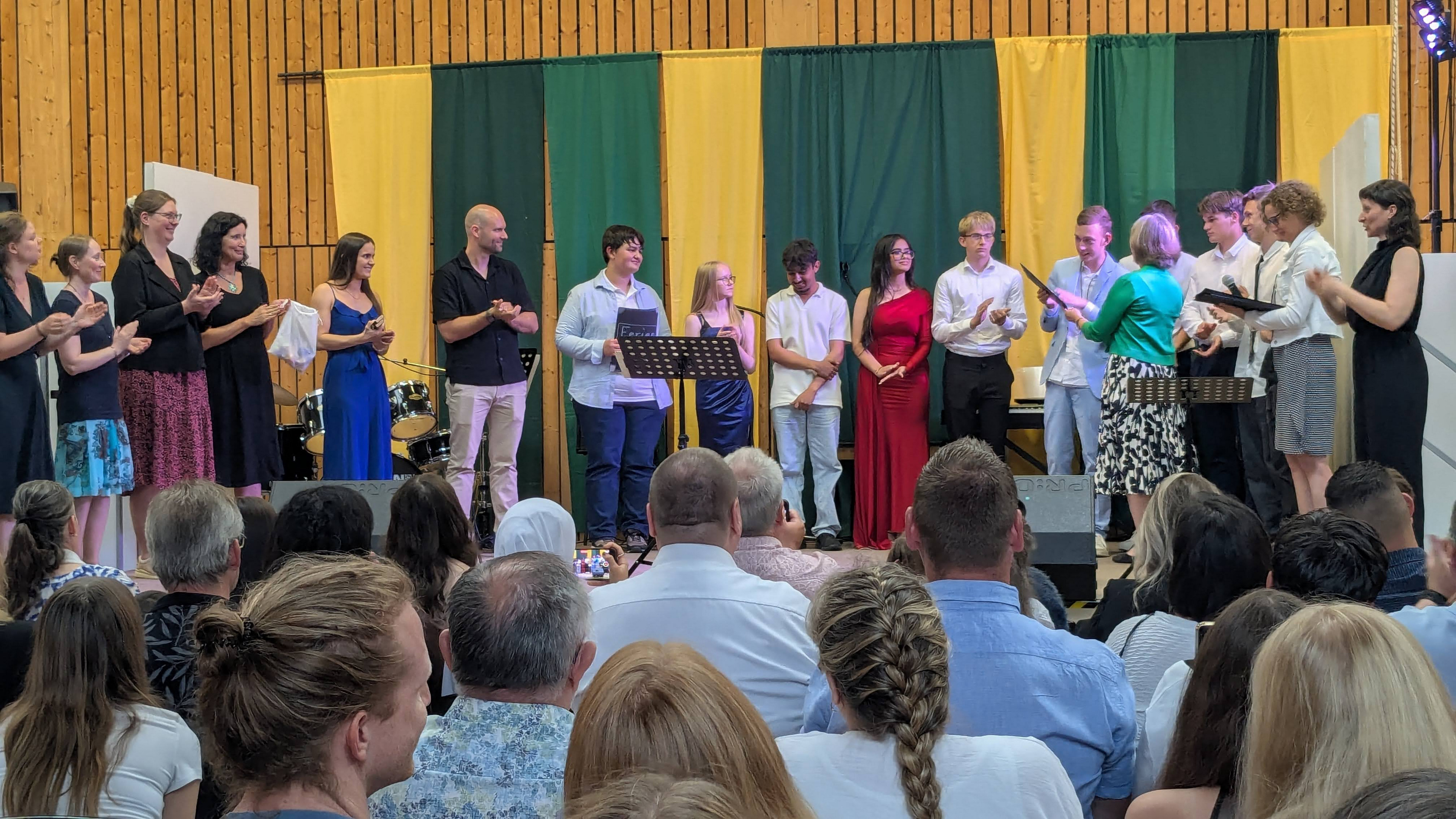 Entlassschülerinnen und Schüler mit Lehrkräften bei der Zeugnisvergabe auf der Bühne. 