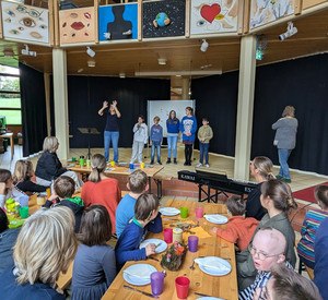 Schüler und Lehrkräfte sind an Tischen im Foyer und eine Klasse mit Schüler steht auf der Bühne und trägt etwas vor. 