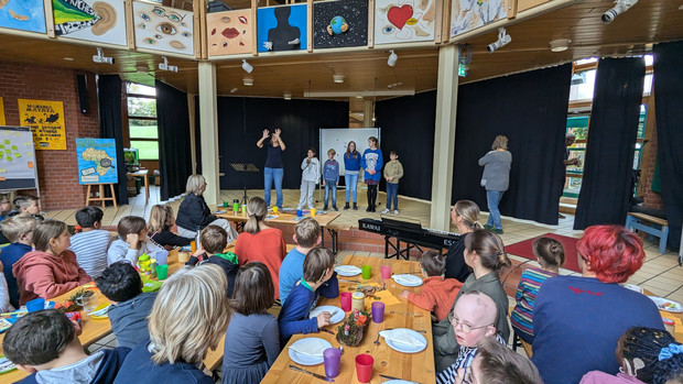 Schüler und Lehrkräfte sind an Tischen im Foyer und eine Klasse mit Schüler steht auf der Bühne und trägt etwas vor. 