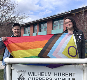 2 Personen halten eine bunte queere Fahne über das Namensschild der Cüppers-Schule.