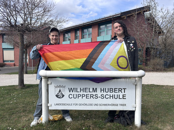 2 Personen halten eine bunte queere Fahne über das Namensschild der Cüppers-Schule. 