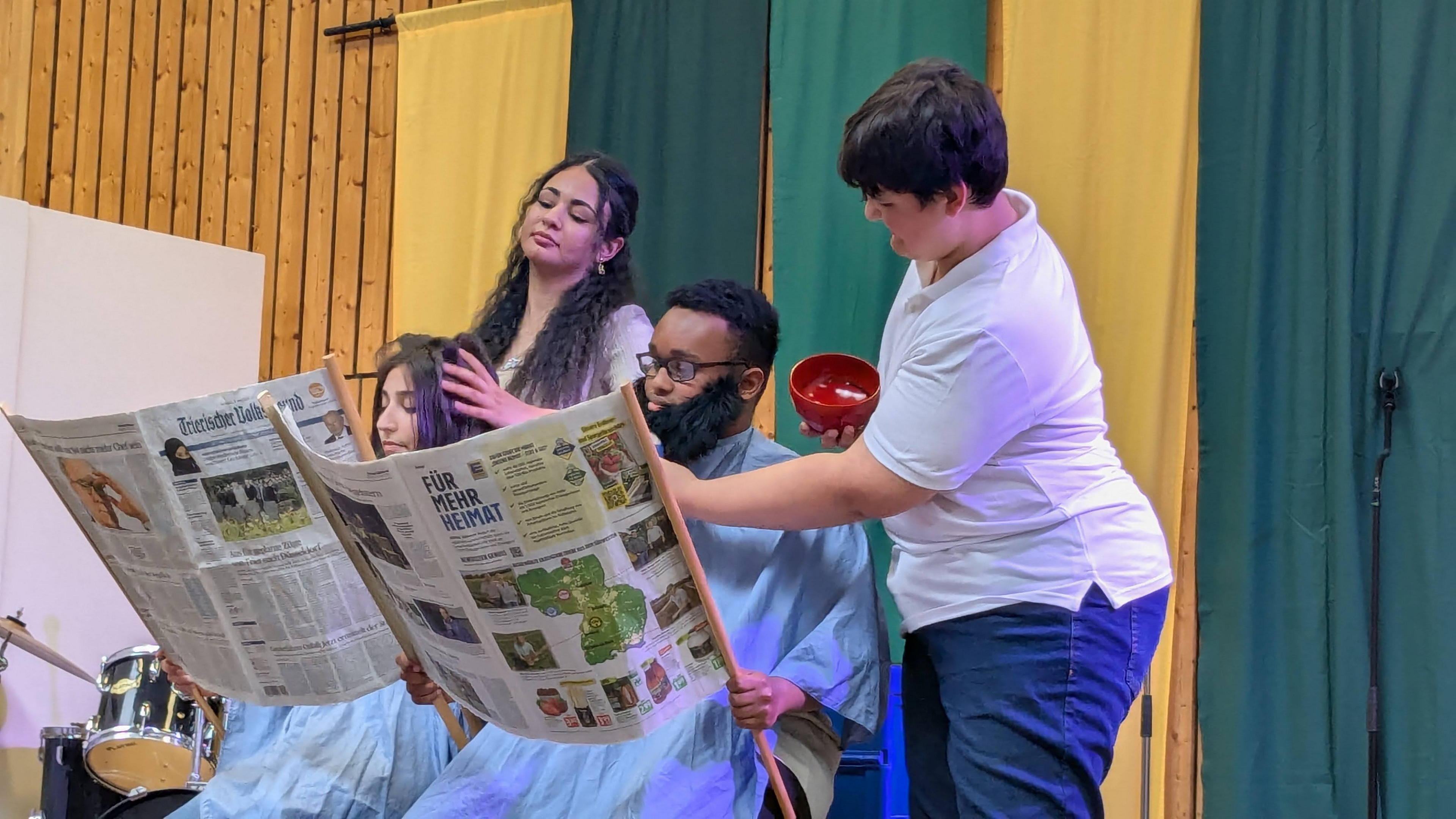 1 Schülerin und 2 Schüler führen ein Sketch auf der Bühne in der Turnhalle. 